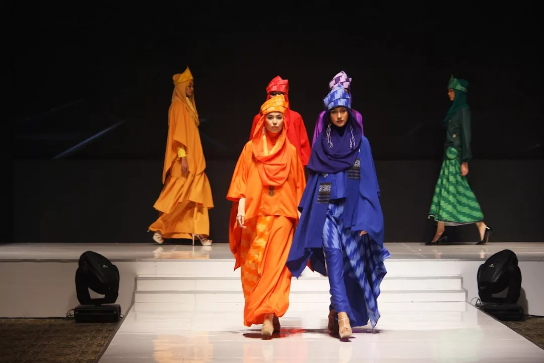 Models walk a runway for Glamesia, wearing brightly colored, modest fashion outfits in orange, red, blue, and green tones with matching head coverings; the background is dark for a touch of luxury.
