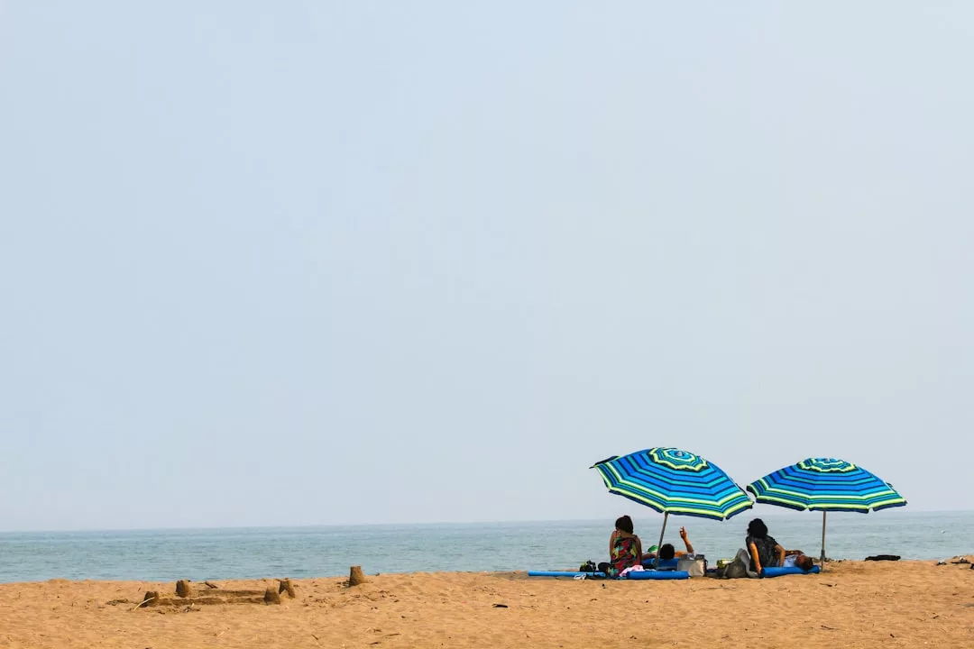 Three people enjoy a stylish lifestyle under two blue-striped umbrellas on a sandy beach, facing the sea beneath a clear sky.