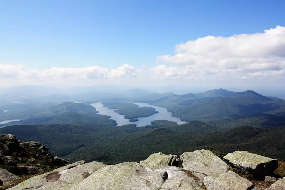 Rocky foreground overlooking a forested landscape with a winding lake and distant mountains under a partly cloudy sky, offering a glimpse into the luxury lifestyle of nature’s finest views.