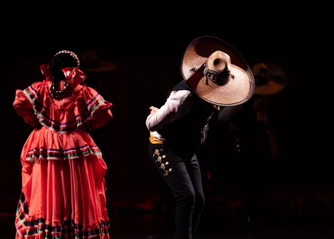 Two dancers in traditional Mexican attire showcase vibrant lifestyle and fashion on stage; one wears a large sombrero and the other dazzles in a red folkloric dress, embodying the spirit of Glamesia.