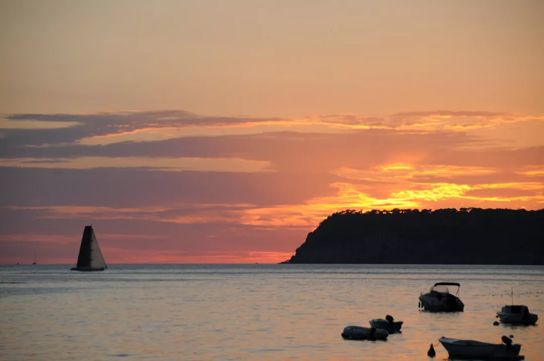 A sailboat and several small boats float on calm water at sunset, with an island silhouette and orange clouds in the background, capturing the essence of a luxury lifestyle.