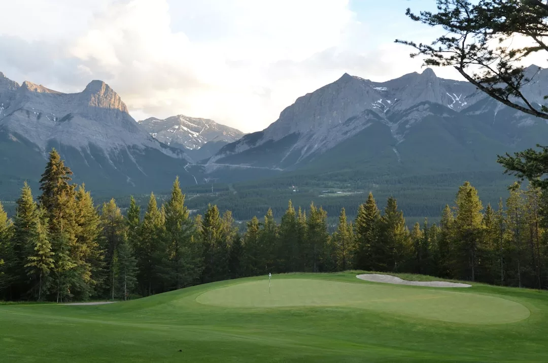 A luxury golf green with a sand bunker in the foreground, dense pine trees in the midground, and tall, rocky mountains in the background under a partly cloudy sky.