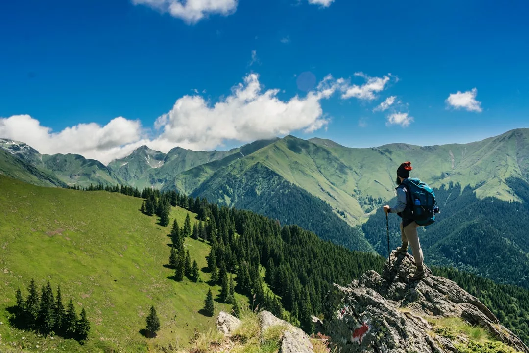 A hiker with a backpack and trekking pole stands on a rocky outcrop, savoring the Glamesia lifestyle while overlooking green mountains and a valley under a blue sky with scattered clouds.