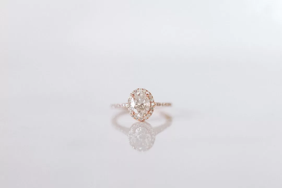 A rose gold Glamesia ring with an oval-shaped clear gemstone is displayed on a reflective white surface, exuding modern fashion elegance.