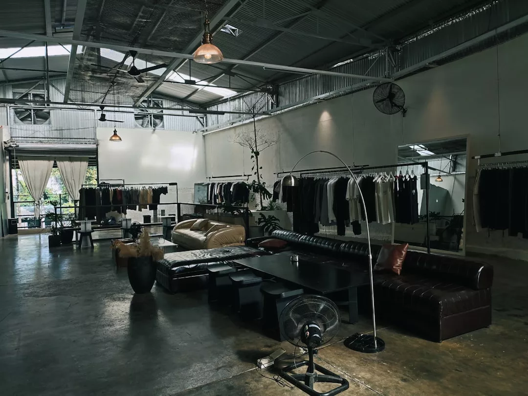 Spacious industrial-style clothing store featuring Fashion-forward racks, leather sofas, a coffee table, and large windows that fill the space with natural light. High ceilings add to the chic ambiance.