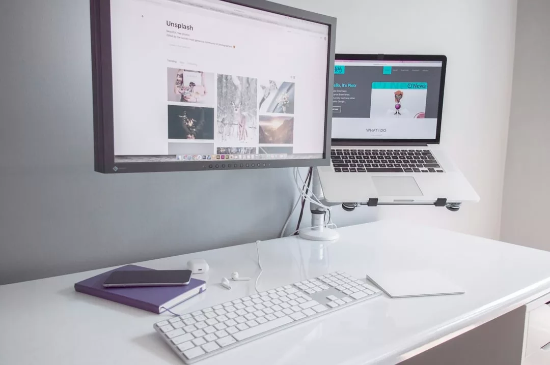 A minimalist luxury white desk setup with a monitor, laptop on a stand, wireless keyboard, mouse, notebook, and earphones arranged neatly.