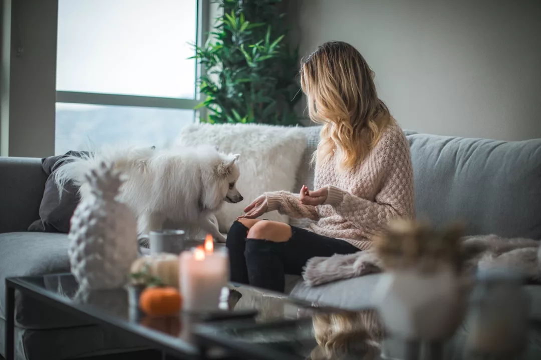 A woman in a beige sweater sits on a gray couch, holding out her hand to a fluffy white dog. Candles and plants on the coffee table complete this cozy luxury lifestyle scene.
