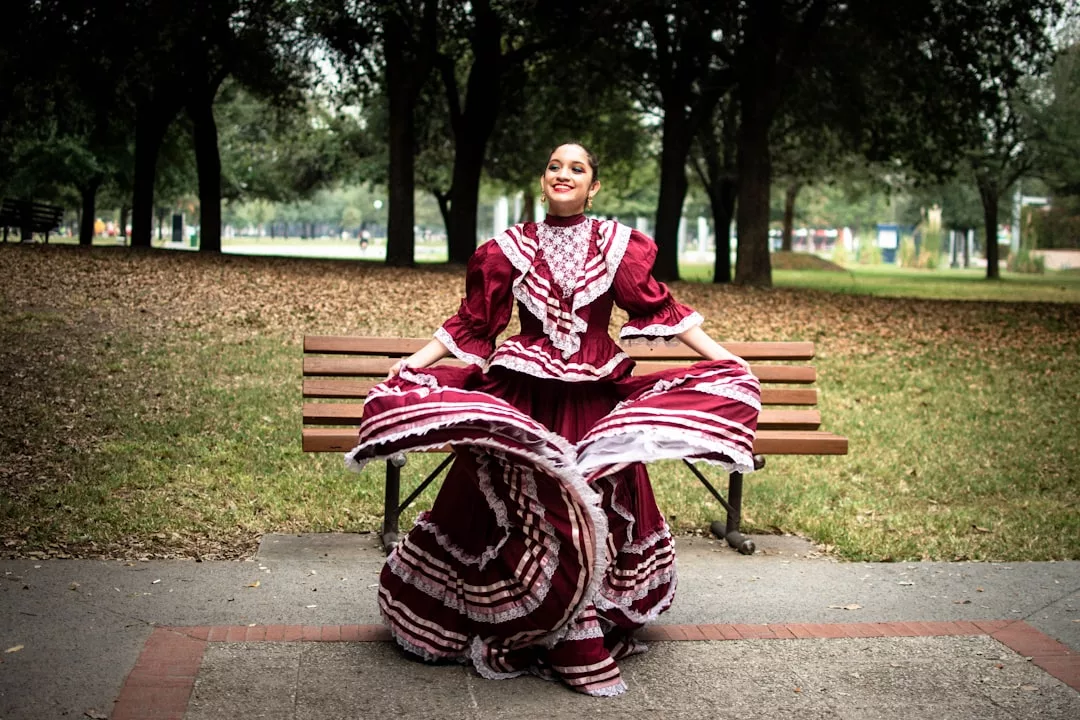 A woman in a traditional maroon and white folkloric dress showcases her fashion sense, holding out her skirt with both hands in front of a park bench. This Glamesia lifestyle moment unfolds with trees and grass in the background.