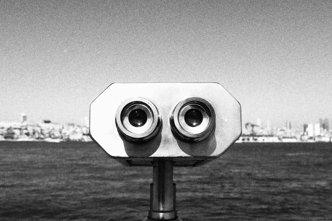 A metal coin-operated binocular viewer faces the camera with water and a luxury cityscape blurred in the background.