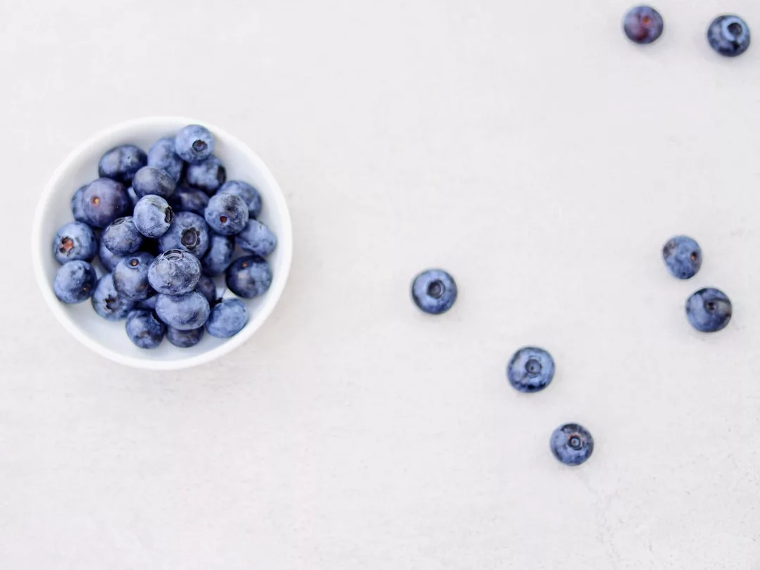 A small white bowl filled with blueberries sits on a light surface, with several loose blueberries scattered nearby—an effortless touch of luxury in everyday living.