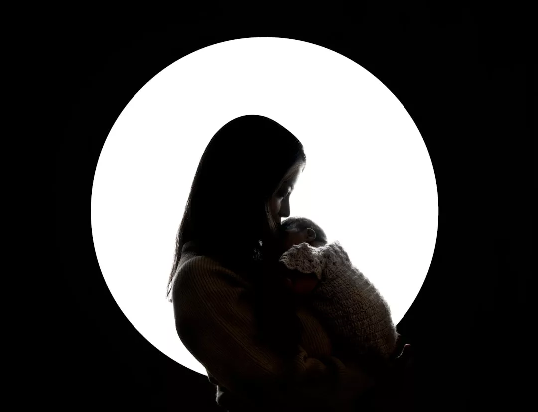 Silhouette of a person holding a baby in front of a large white circular light source against a dark background, capturing the essence of luxury lifestyle and Glamesia's signature elegant aesthetic.