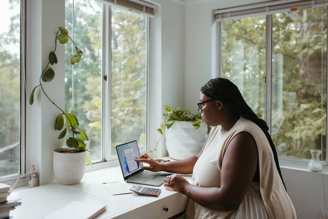 A person sits at a desk by a window, embracing a modern lifestyle while using a calculator and looking at a laptop displaying a chart or graph. Fashionable potted plants decorate the windowsill.