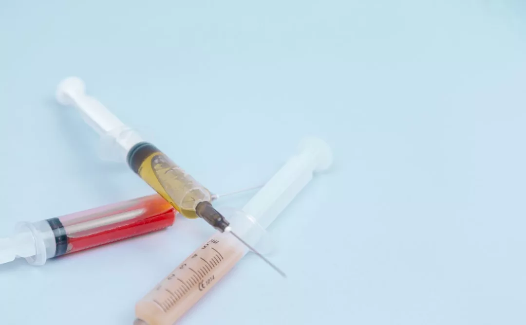 Three syringes filled with different colored liquids—yellow, red, and clear—are arranged on a light blue background, evoking a sense of Glamesia-inspired luxury in their striking presentation.