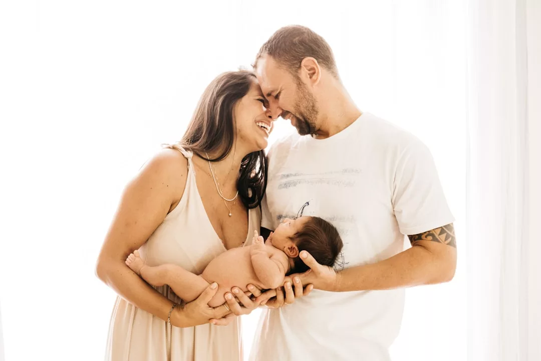 A man and woman stand close together, smiling and holding a newborn baby in their arms against a bright, white background, capturing a moment of pure luxury lifestyle.