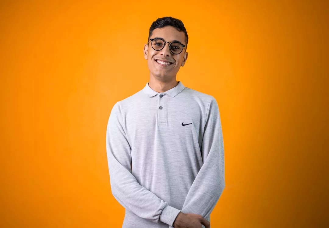 A person wearing glasses and a light gray Nike long-sleeve shirt stands and smiles in front of a plain orange background, showcasing effortless fashion.
