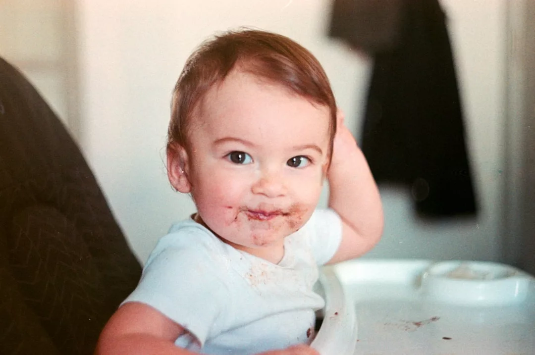 A baby sitting in a high chair with food smeared around their mouth, enjoying a playful moment that captures the essence of family lifestyle, looking at the camera.