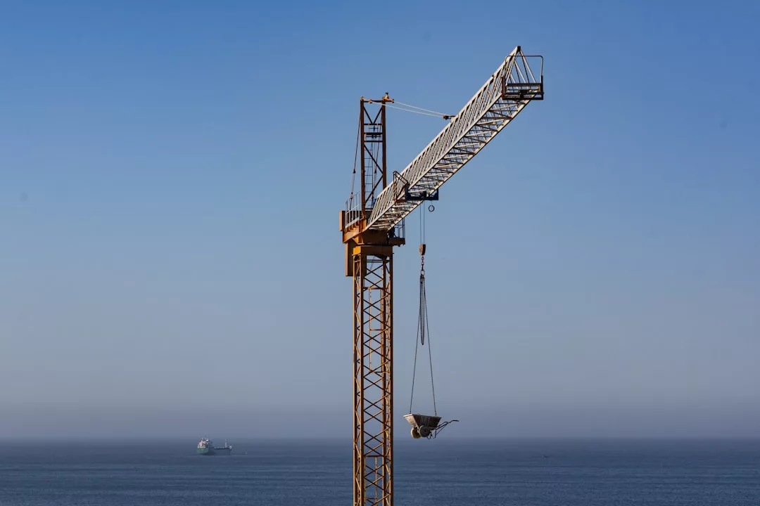 A construction crane with a suspended load stands near the ocean, with a ship visible on the water in the background under a clear blue sky, adding a touch of Glamesia to this coastal lifestyle scene.