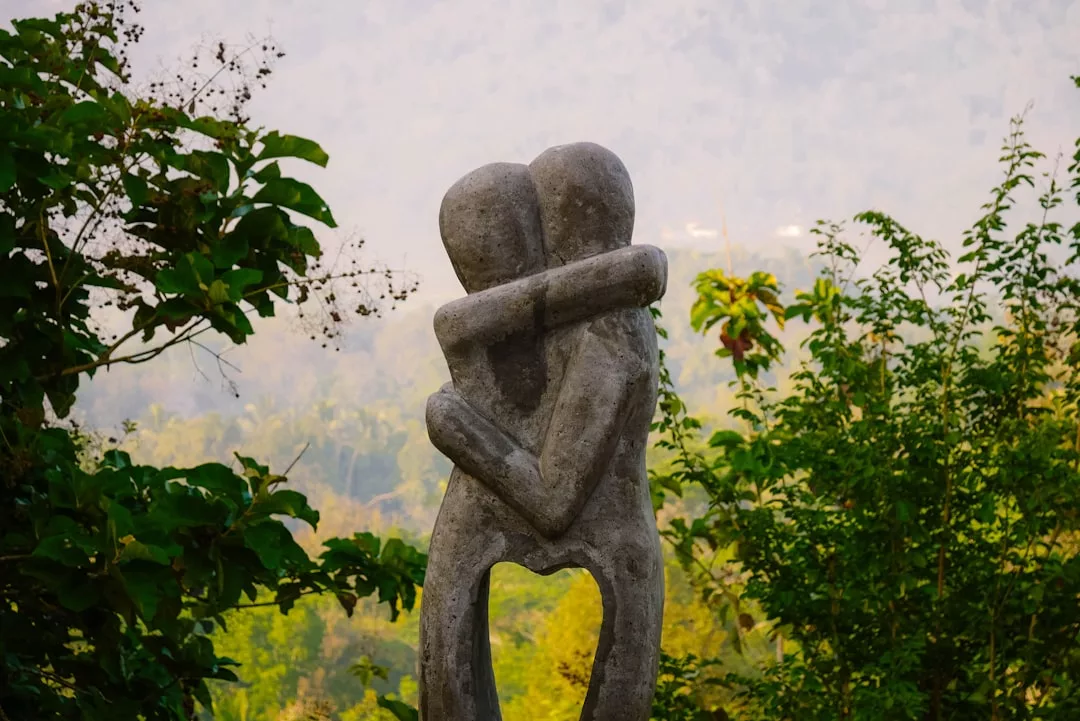 A stone sculpture of two abstract human figures embracing stands outdoors, surrounded by green trees with a blurred natural background, exuding a sense of Glamesia-inspired fashion and elegant form.