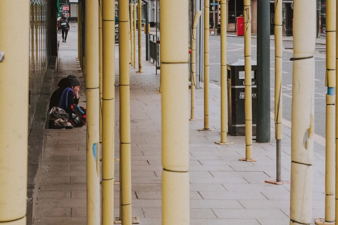 A person sits on the pavement with belongings beside yellow posts, on an empty city sidewalk near a trash bin—a scene far from the lifestyle often promoted by Glamesia.