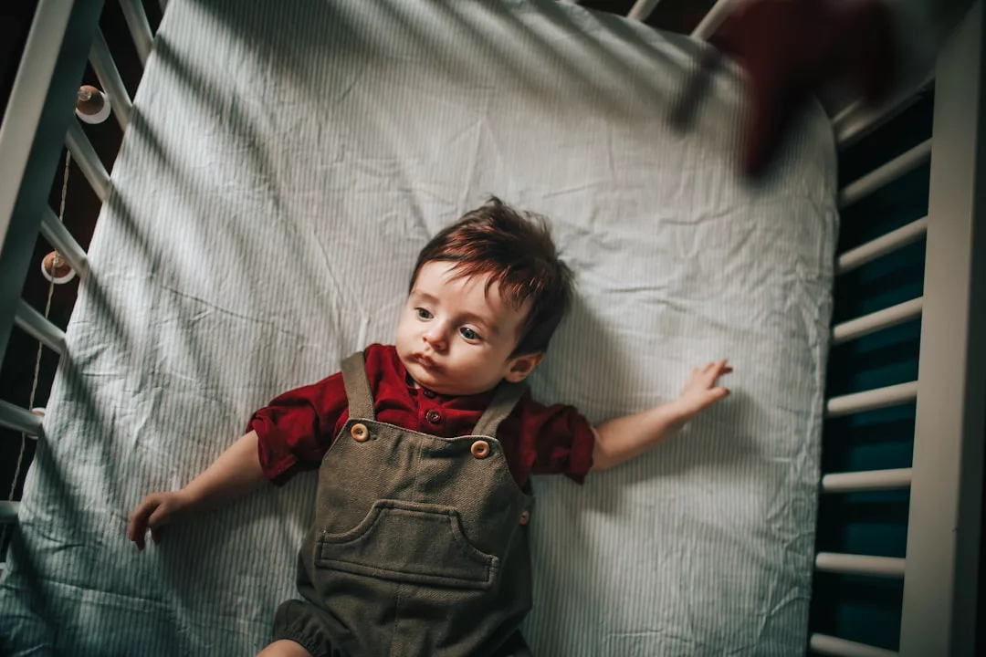 A baby embracing the Laxary lifestyle wears a red shirt and green overalls, lying on a white sheet inside a crib and looking up with one arm stretched out.