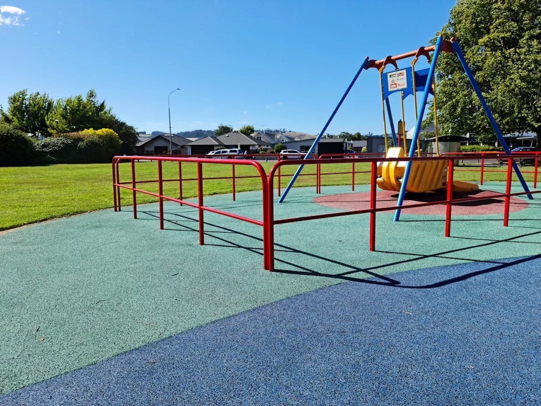 A playground with a red metal fence, blue and green rubber flooring, and a swing set with a boat-shaped seat under a clear blue sky offers a vibrant outdoor lifestyle for families.