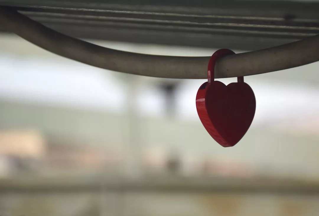 A red heart-shaped padlock hangs on a metal railing, embodying a touch of lifestyle charm against a blurred background.