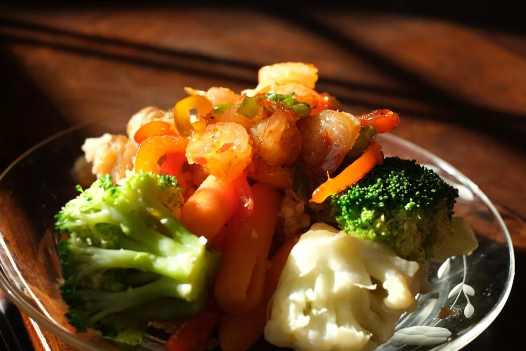 A plate of cooked shrimp, carrots, bell peppers, broccoli, and cauliflower is artfully arranged on a glass dish, bathed in natural sunlight for a touch of luxury lifestyle dining.