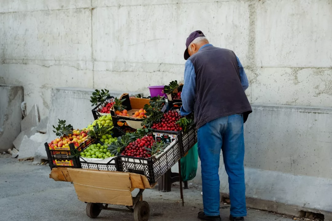 An older man arranges fruit on a street cart, which holds crates filled with strawberries, grapes, tomatoes, and other produce, reflecting a simple lifestyle against a concrete wall.