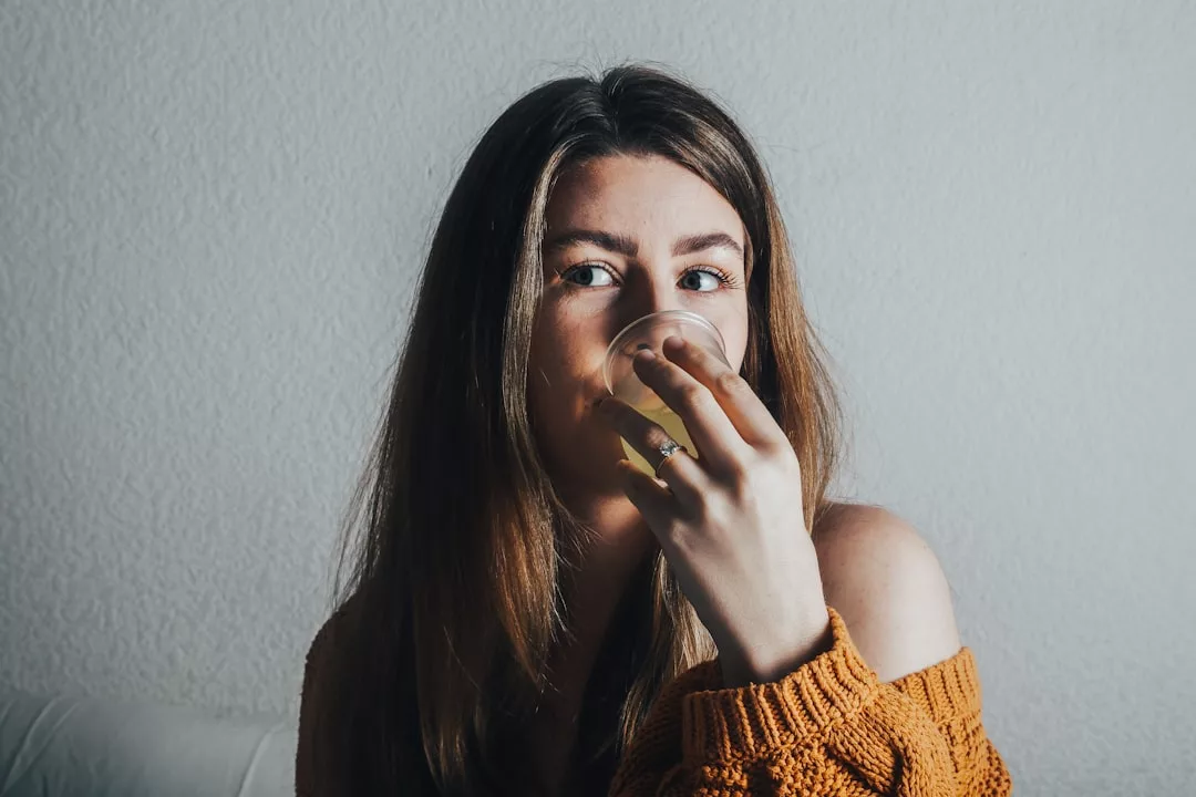 A woman with long brown hair wearing a luxury orange sweater sits indoors, holding a glass up to her mouth and looking slightly to the side.
