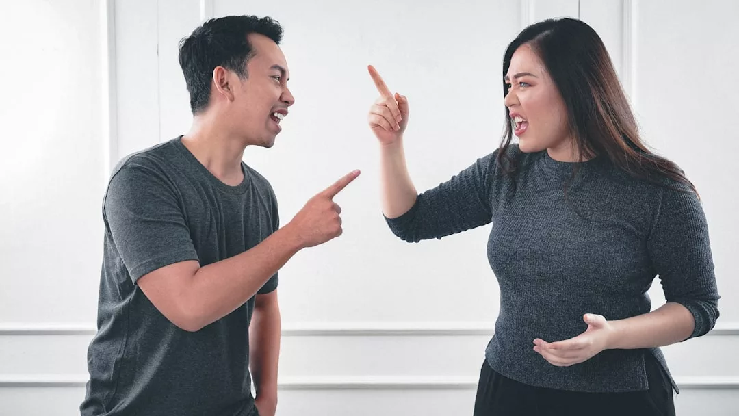 A man and a woman argue indoors, facing each other and pointing fingers while expressing strong emotions in a setting of understated luxury.