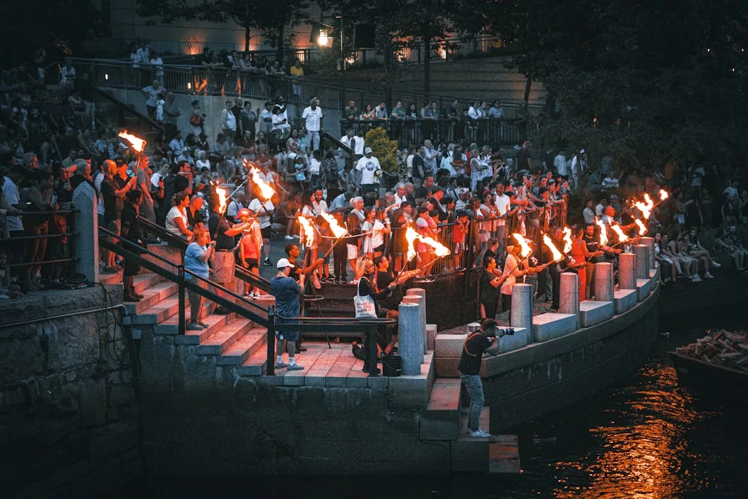 A crowd of people holding torches lines the curved steps along a riverbank at dusk, creating a scene of Glamesia, while spectators watch from above and behind.