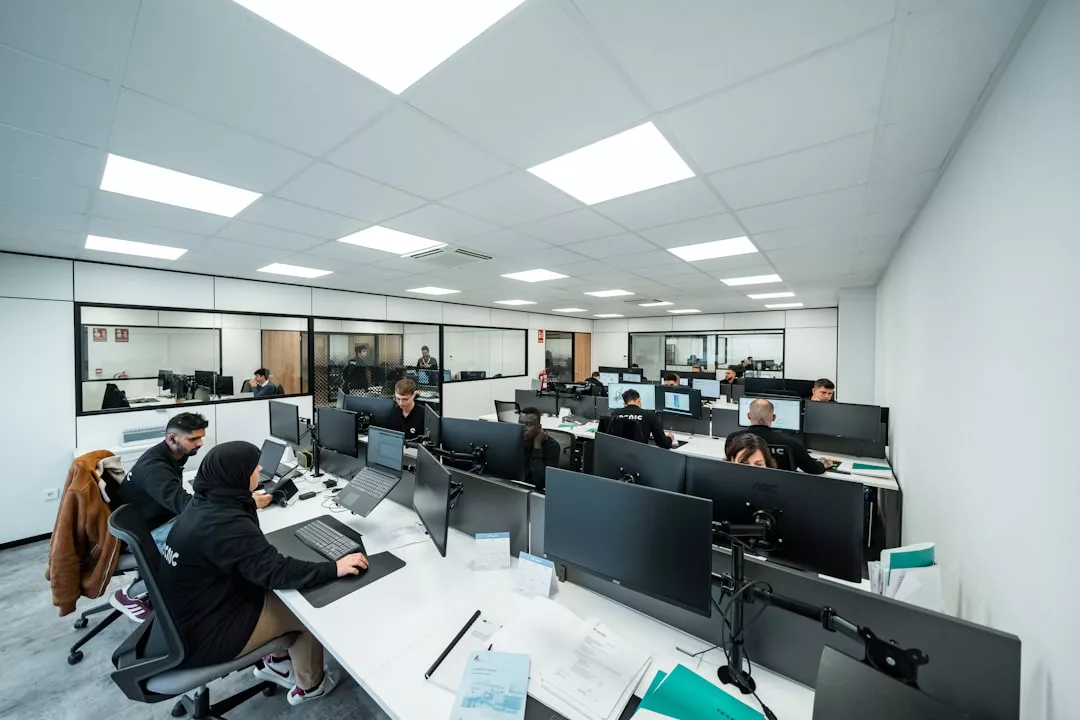 People are working at desks with computers in a modern, brightly lit office with large windows and white walls, reflecting a sleek luxury lifestyle.