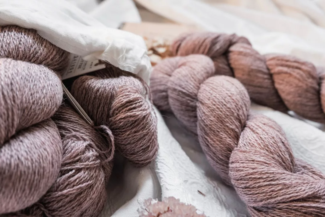Skeins of light mauve yarn and partially unraveled balls of yarn rest on a white textured fabric, adding a touch of Glamesia-inspired lifestyle to your fashion projects.