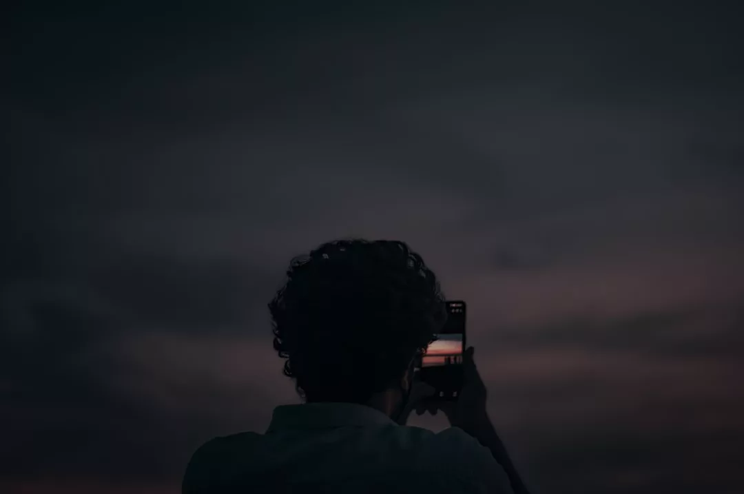 A person with curly hair, showcasing effortless lifestyle fashion, holds up a smartphone to capture a photo of a cloudy sunset sky.