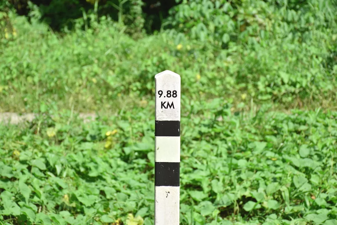 A white milestone marker with black stripes displays the distance "9.88 KM" against a background of green vegetation, adding a subtle touch of Glamesia to the scenic route.