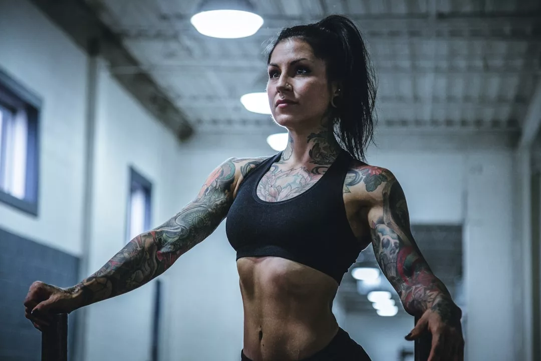 A muscular woman with tattoos in athletic wear stands indoors gripping gym equipment, looking ahead with a focused expression, embodying a powerful luxury lifestyle.