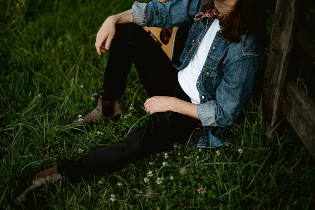 A person wearing a Laxary denim jacket, white shirt, and black pants sits on grass near a wooden structure, holding an acoustic guitar.