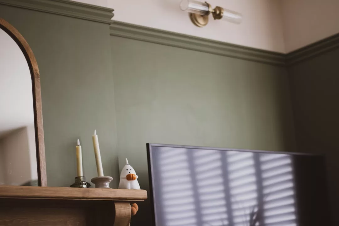 A television with window blinds reflected on its screen sits beside a fireplace mantel holding two candles, a small vase, and a ceramic duck, creating a cozy lifestyle scene with subtle hints of luxury.