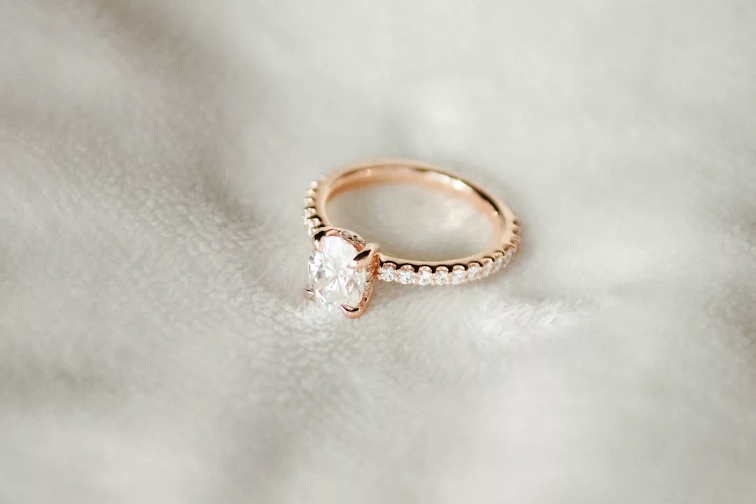 A Glamesia gold ring with a large oval-cut clear gemstone and small diamonds on the band, resting on a soft, white fabric surface—perfect for expressing luxury and timeless fashion.