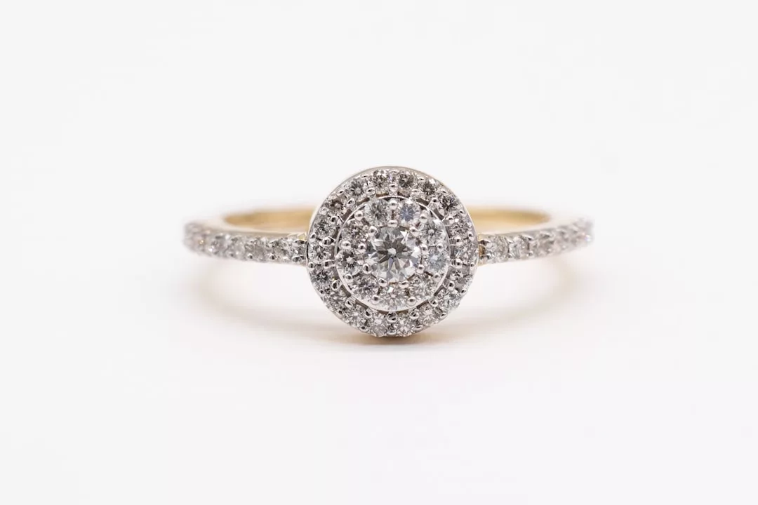 A luxury diamond ring with a round center stone, encircled by a double halo of smaller diamonds and a diamond-set band, photographed on a white background.