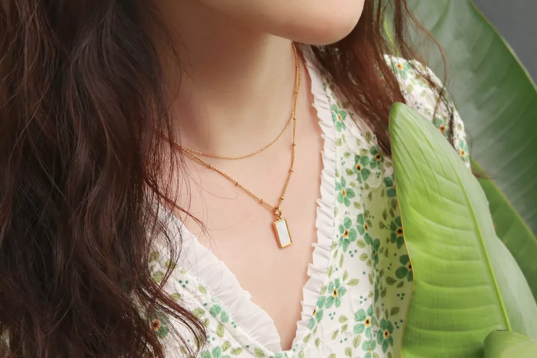 A woman wearing a floral dress and two gold necklaces, one with a rectangular pendant, stands next to a large green leaf, radiating Glamesia Fashion charm.