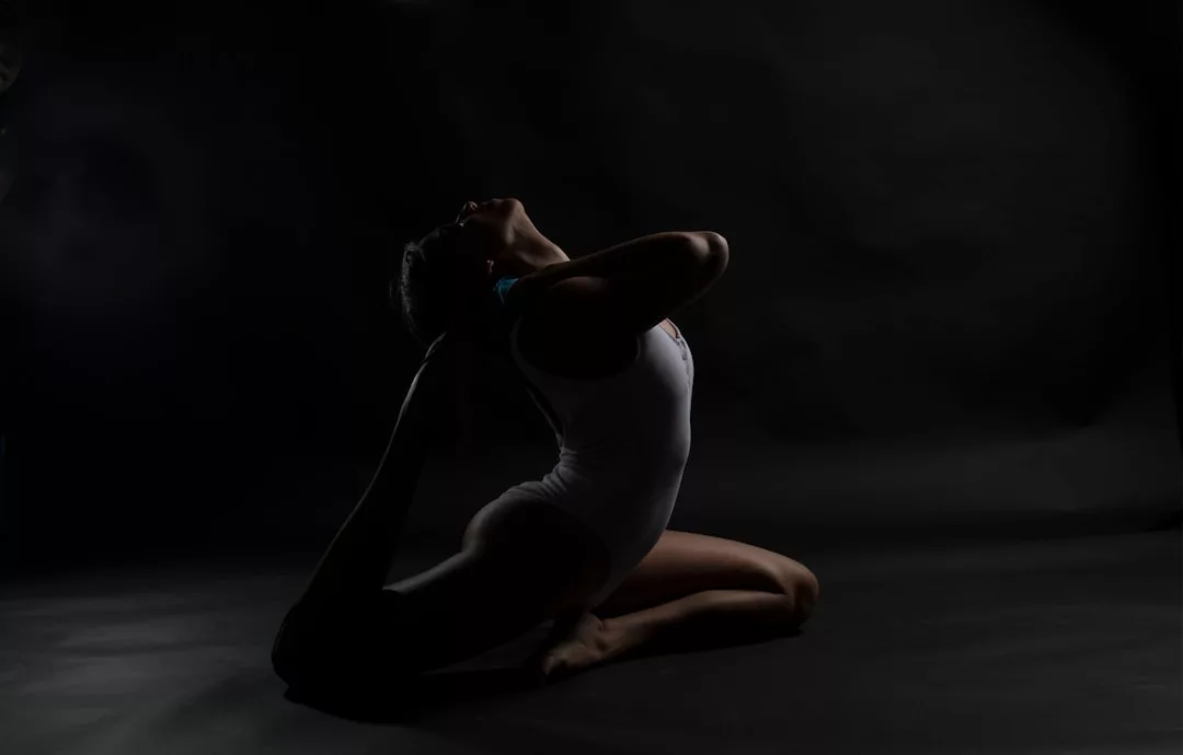 Person in a white bodysuit performing a yoga pose on the floor, arching their back and holding one foot with both hands in a dark, low-light setting, exuding an aura of Laxary and refined grace.