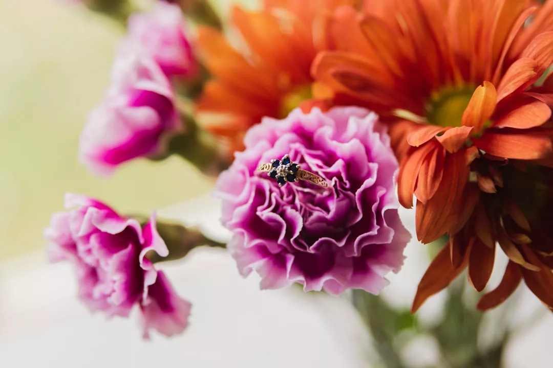 A gold ring with a dark blue gemstone rests on a pink carnation flower, evoking luxury, with orange and pink flowers visible in the background.