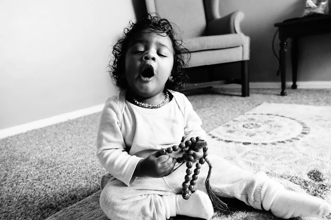A young child sits on a carpeted floor holding prayer beads, eyes closed and mouth open, in a living room adorned with touches of luxury—a stylish chair and table enhancing the elegant setting.