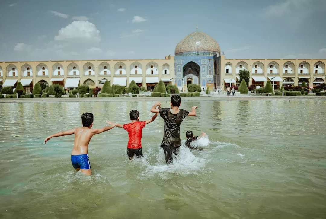 Four boys embrace a carefree lifestyle as they run into a shallow pool in front of the Shah Mosque, its blue tiled dome and arched facade glowing under the sun.