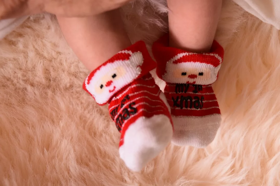 A baby's feet wearing red and white Christmas-themed socks with Santa faces, resting on a soft, beige surface—a charming touch of festive fashion for your little one's cozy lifestyle.