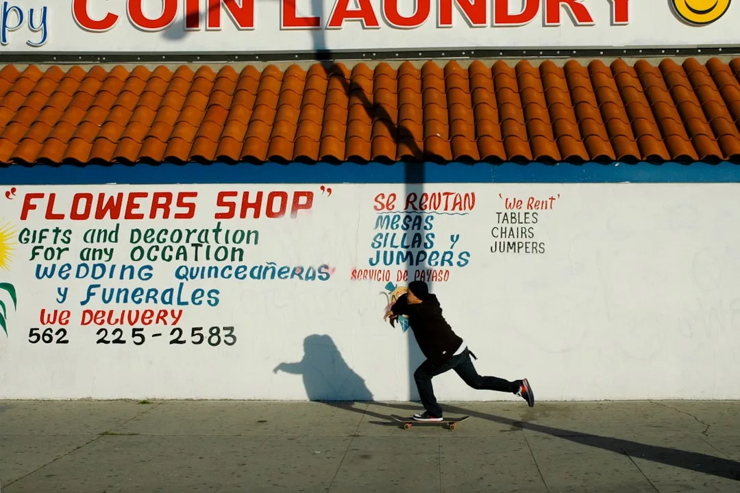 A person in dark clothing rides a skateboard past a wall with colorful painted advertisements for a flower shop and party rentals beneath a "Coin Laundry" sign, blending street style with Glamesia fashion vibes.