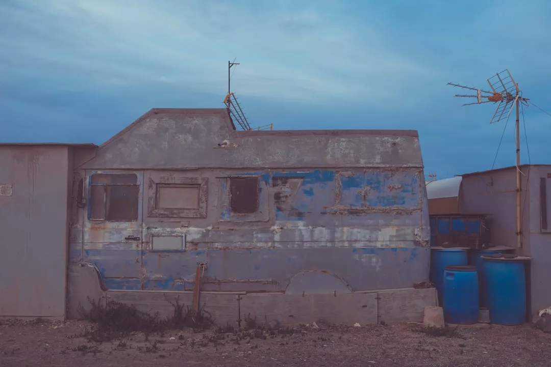 A weathered, blue and gray camper sits between two structures, with antennas on top and blue barrels nearby, hinting at a unique lifestyle under a cloudy sky.