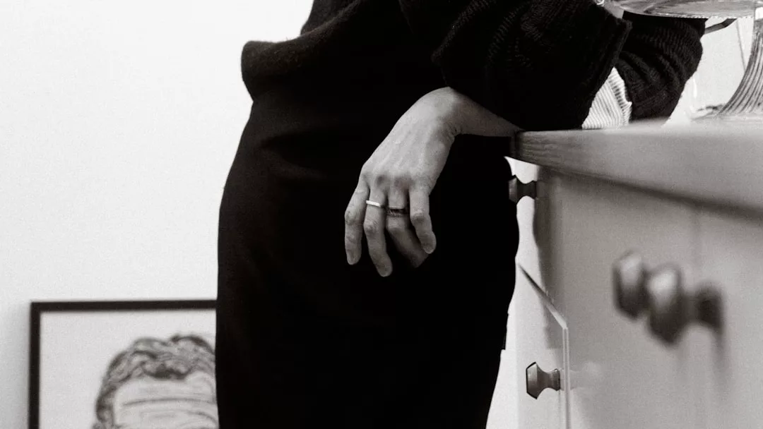 A person in dark clothing leans against a kitchen counter, their left hand resting on the edge; a framed sketch is partially visible in the background, adding a touch of Fashion to the stylish scene.