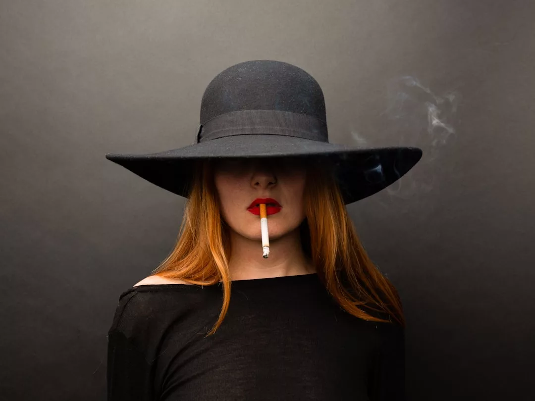 Exuding Glamesia luxury, a woman with long red hair dons a wide-brimmed black hat and top, holding a lit cigarette between her lips; her eyes remain hidden beneath the hat, embodying an enigmatic lifestyle.